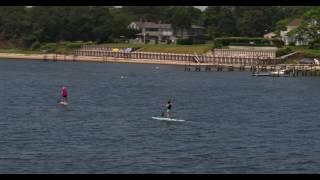 Low aerial tracking shot of two female paddleboarders, with beachhouses in the background.